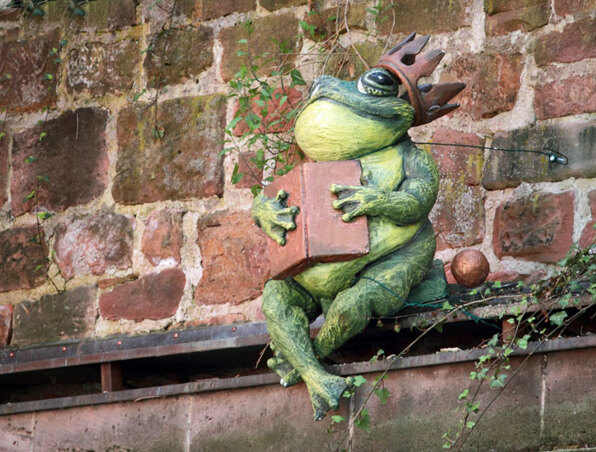 Dekorative Skulptur eines grünen Frosches mit Krone, sitzend auf einem Sims vor einer alten Steinmauer mit Efeu