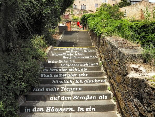 Historisches Schloss über einer Steintreppe mit weißem Zitat auf den Stufen, umgeben von üppigem Grün auf beiden Seiten