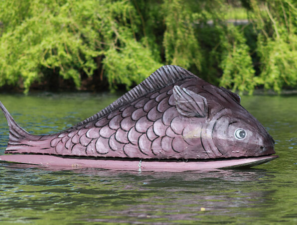 Große dekorative Fischskulptur schwimmt auf ruhigem Wasser, umgeben von üppigem Grün für eine entspannte Außenatmosphäre
