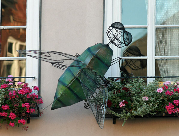 Große dekorative Metallinsekten-Skulptur an Hotelzimmerfenster mit lebhaften rosa und roten Blumen im Blumenkasten