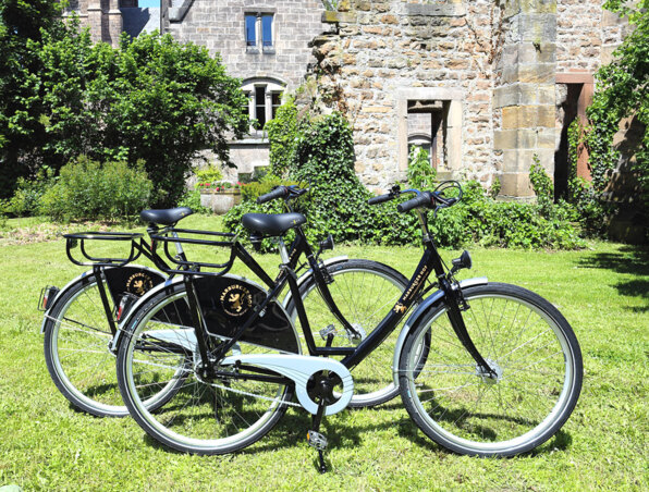 Zwei schwarze Gäste-Fahrräder auf grüner Wiese neben einem historischen Steinbau mit Kletterpflanzen
