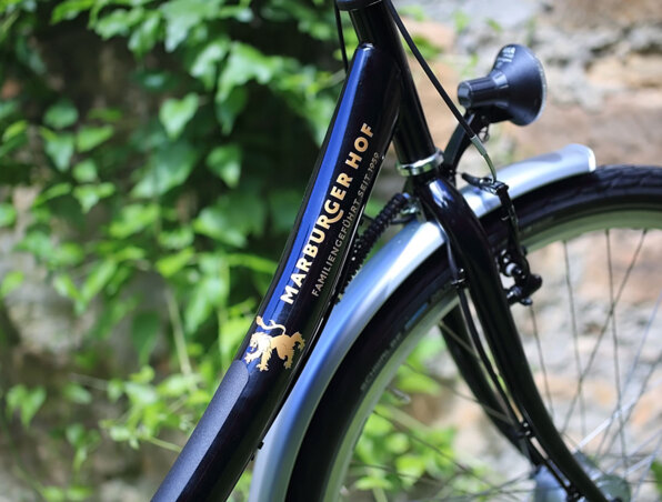 Nahaufnahme eines schwarzen Fahrradrahmens mit Marburger Hof Logo, im Freien neben Grünpflanzen und Steinmauer geparkt