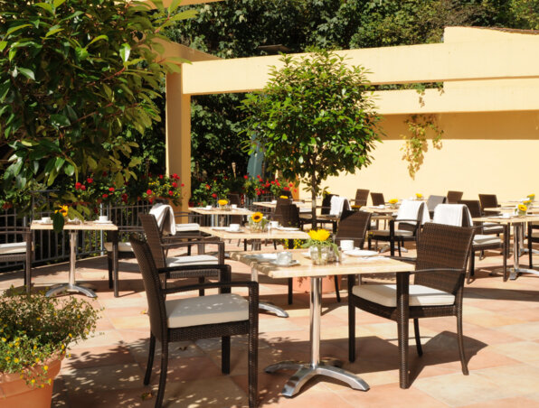 Sonnige Außenterrasse des Hotels mit Korbsesseln, Marmortischen, Topfpflanzen und kleinen gelben Blumenarrangements