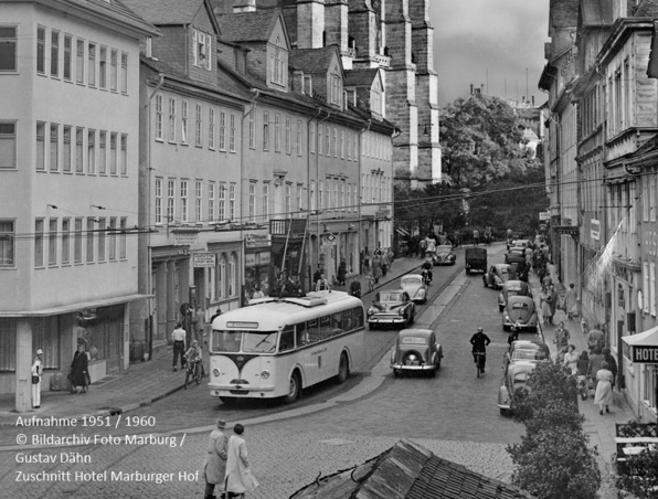 Historische europäische Straße mit Oldtimerautos, einem klassischen Bus, Fußgängern und einem Hotelschild an Kopfsteinpflaster