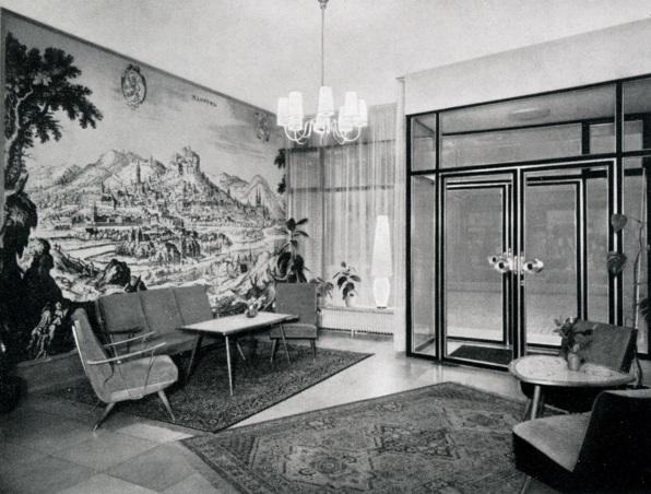 Hotellobby mit modernen Sitzmöbeln, dekorativen Teppichen, großem Wandgemälde einer Berglandschaft, Glastüren und stimmungsvoller Beleuchtung.