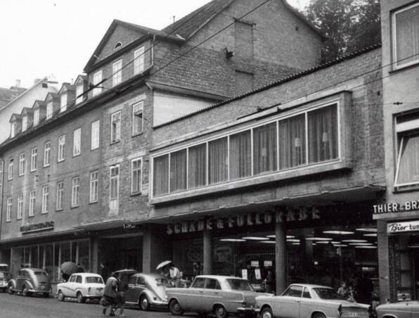 Historisches Hotelgebäude mit klassischer Architektur und Geschäften im Erdgeschoss, Oldtimer-Autos auf der Straße, Fußgänger mit Regenschirmen