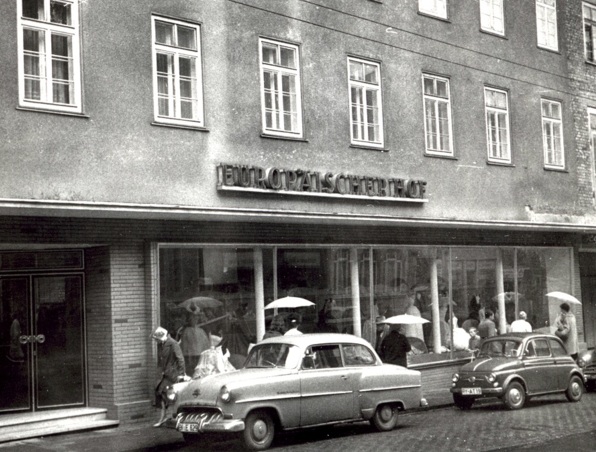 Historischer Hoteleingang mit Oldtimern davor und Gästen mit Regenschirmen an einem Regentag