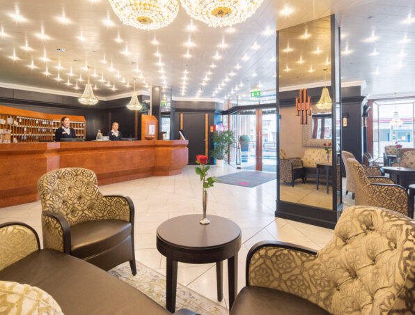Heller Hotellobby mit poliertem Holzeingangstresen, freundlichem Personal, eleganten Sesseln, Kronleuchtern und frischen Rosen auf den Tischen