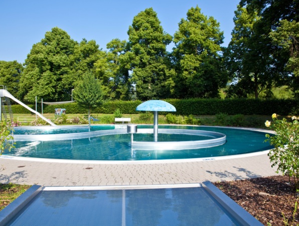 Runder Außenpool mit Wasserrutsche und Pilzbrunnen, umgeben von Bäumen und gepflasterter Terrasse bei Sonnenschein