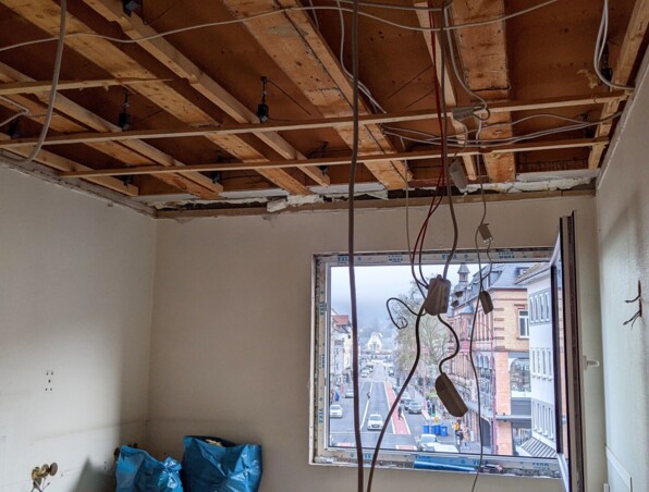 Room under renovation with exposed wooden ceiling beams, hanging electrical wires, and a street view through an open window