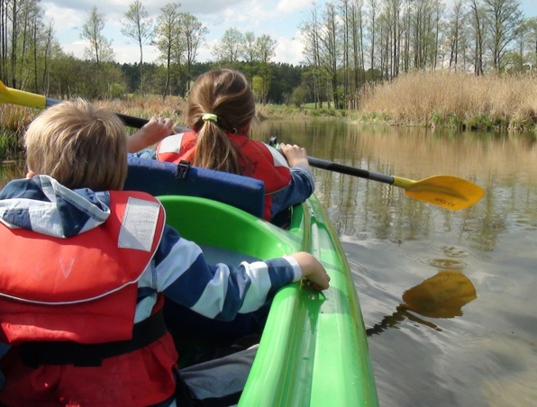 Zwei Kinder in Schwimmwesten paddeln auf einem ruhigen Fluss, umgeben von hohem Gras und Bäumen bei leicht bewölktem Himmel