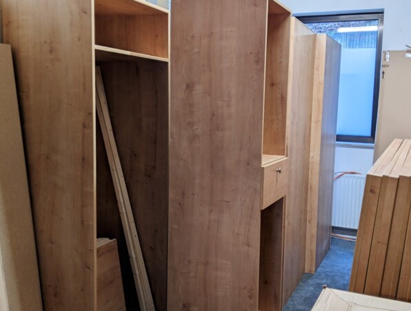 Unassembled wooden furniture panels and cabinets stacked in a storage room with a small window and carpeted floor