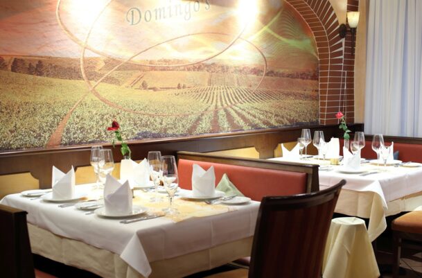 Elegant restaurant seating with white tablecloths, folded napkins, wine glasses, and a mural of vineyard scenery