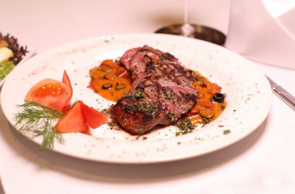 Grilled steak slices with herb garnish and savory tomato-olive sauce, served with fresh tomato wedges and dill on a white plate
