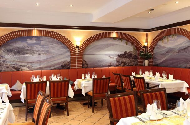Elegant hotel dining area with red leather banquette, striped wooden chairs, white tablecloths, and coastal mural accents on arched walls