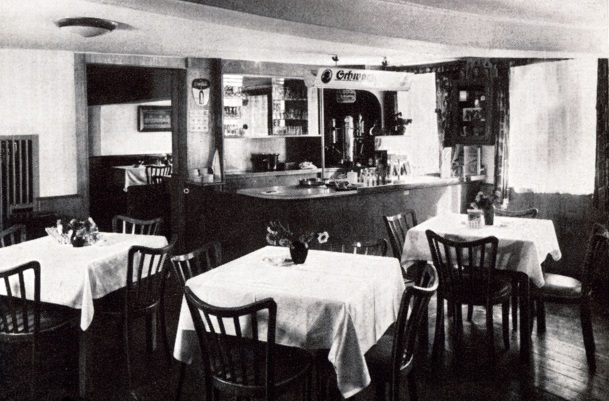 Gemütlicher Speiseraum im Hotel mit Holzstühlen, weißen Tischdecken, frischen Blumenarrangements und kleiner Bar im Hintergrund