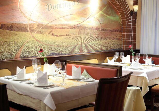 Elegant restaurant seating with white tablecloths, folded napkins, wine glasses, and a mural of vineyard scenery