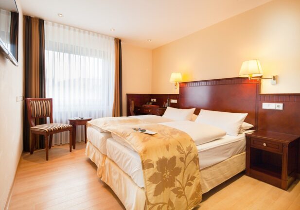 Bright hotel room with twin beds, elegant wooden headboard, striped chair, and sheer curtains letting in natural light