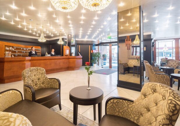 Bright hotel lobby with polished wooden reception desk, elegant chandeliers, patterned armchairs, and fresh red roses on tables
