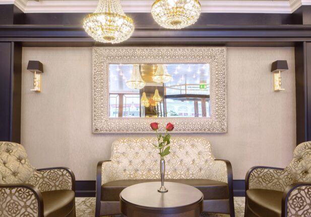 Elegant hotel lounge with patterned armchairs, tufted loveseat, ornate mirror, crystal chandeliers, and vase with two red roses on table