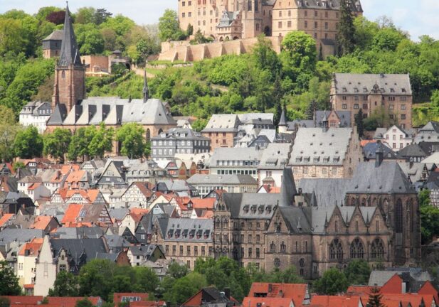 Historic castle atop green hill overlooking red-roofed town with Gothic church steeples and lush trees