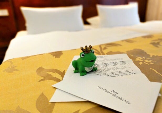 Hotelbett mit weißen Kissen, gelber Bettdecke mit Blumenmuster und kleinem grünen Frosch mit Krone auf Willkommensbrief