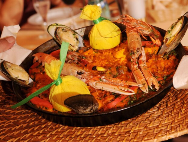 Traditionelle Meeresfrüchte-Paella mit Garnelen, Miesmuscheln und Zitronen im Stoffbeutel in schwarzer Pfanne auf geflochtenem Untersetzer