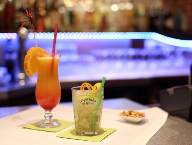 Zwei farbenfrohe Cocktails mit Fruchtgarnitur auf einer Bar, Schale mit Nüssen und blauem LED-Licht im Hintergrund