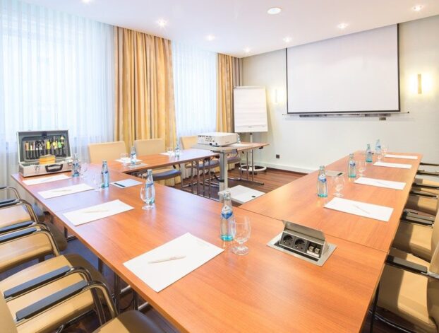 Heller Konferenzraum mit U-förmigen Holztischen, beigen Lederstühlen, Wasserflaschen, Notizblöcken, Beamer und viel Tageslicht