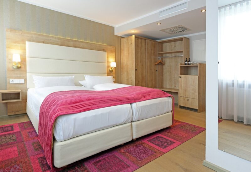 Modernes Hotelzimmer mit zwei zusammengestellten Betten, pinker Tagesdecke, Holzkleiderschrank, weicher Beleuchtung und großem Fenster mit Gardinen