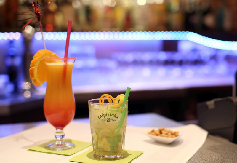 Zwei farbenfrohe Cocktails mit Fruchtgarnitur und Strohhalmen auf einer Bar mit blauer LED-Beleuchtung und einer Schale Nüsse daneben