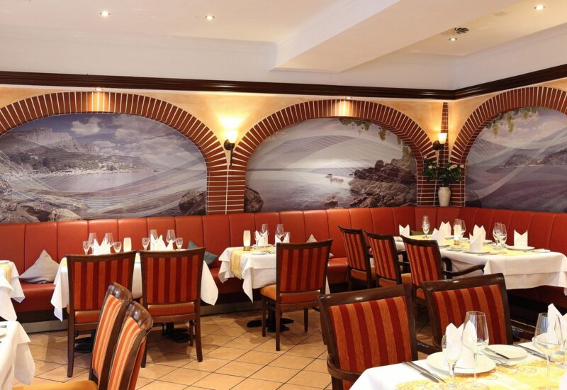 Eleganter hotelrestaurantbereich mit rotem Leder-Sitzbank, gestreiften Holzstühlen, weißen Tischdecken und Küstenmuralen an gewölbten Wänden