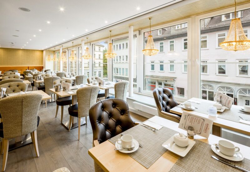 Helles Hotel-Restaurant mit großen Fenstern, eleganten Kronleuchtern, gemusterten und Lederstühlen, Tischen für Frühstück gedeckt.