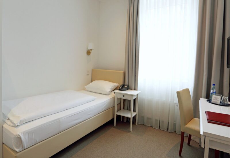 Kompaktes Hotelzimmer mit Einzelbett, Nachttisch mit Telefon, Schreibtisch mit Stuhl, Fernseher und Fenster mit Gardinen