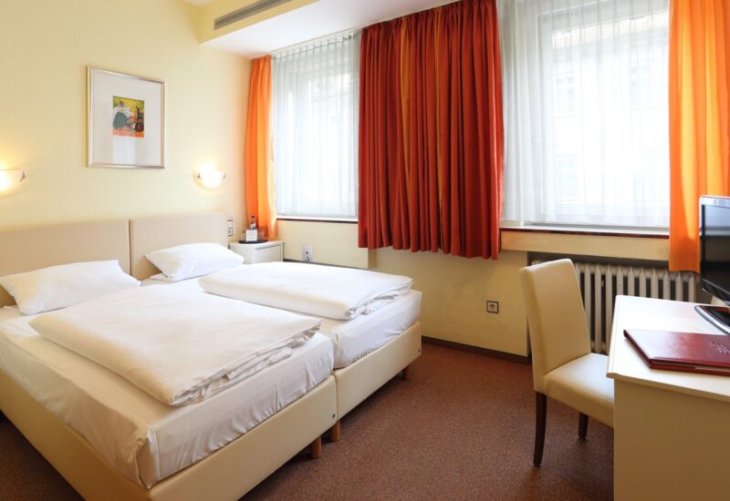 Helles Hotelzimmer mit zwei Einzelbetten, weißer Bettwäsche, orangen Vorhängen, Schreibtisch mit Stuhl und Wandlampen zum Lesen.