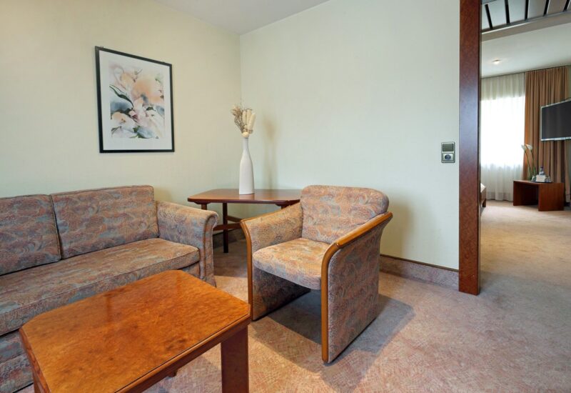 Wohnbereich einer Hotelsuite mit gemustertem Sofa und Sessel, Holztisch, Beistelltisch mit Vase und Tageslicht aus angrenzendem Zimmer