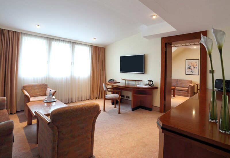 Geräumige Hotelsuite mit Sitzecke, gemusterten Sesseln, Holztisch, Flachbildfernseher und sanftem Tageslicht durch große Fenster