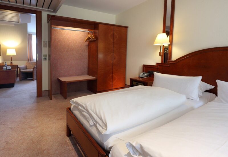 Gemütliches Hotelzimmer mit zwei Einzelbetten, Holz-Kopfteil, Nachttischlampe, offenem Kleiderschrank mit Kleiderbügeln und Sitzbereich mit Sessel