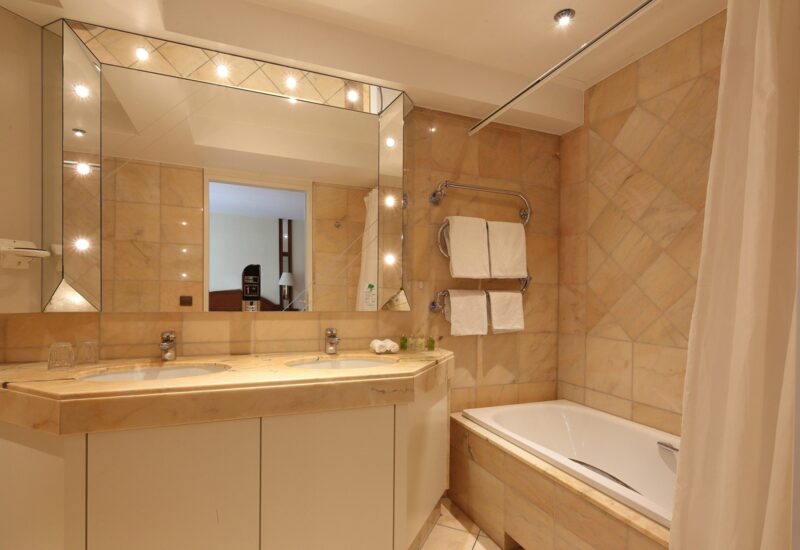 Hotelbadezimmer mit Doppelwaschbecken, großem beleuchtetem Spiegel, beige gefliesten Wänden, Badewanne mit Duschvorhang und ordentlich platzierten Handtüchern und Pflegeprodukten