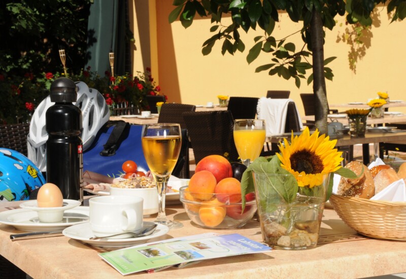 Frühstück im Freien mit frischem Obst, Orangensaft, Kaffee, Brotkorb und Sonnenblume auf sonniger Terrasse mit Gartenbestuhlung
