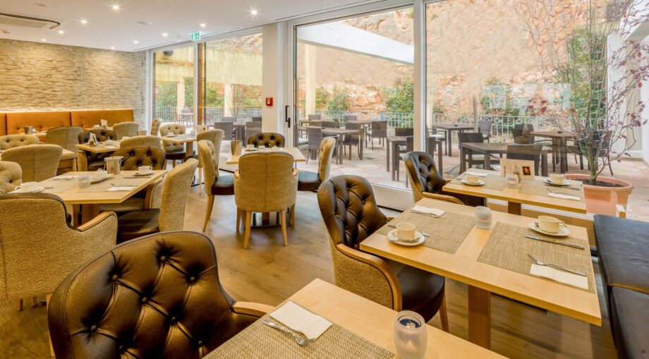 Bright hotel dining area with plush seating, wooden tables set for breakfast, and large glass doors opening to an outdoor terrace.