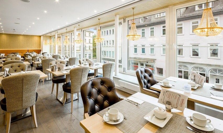 Bright hotel dining room with tufted chairs, large windows overlooking city buildings, and elegant chandeliers hanging from ceiling