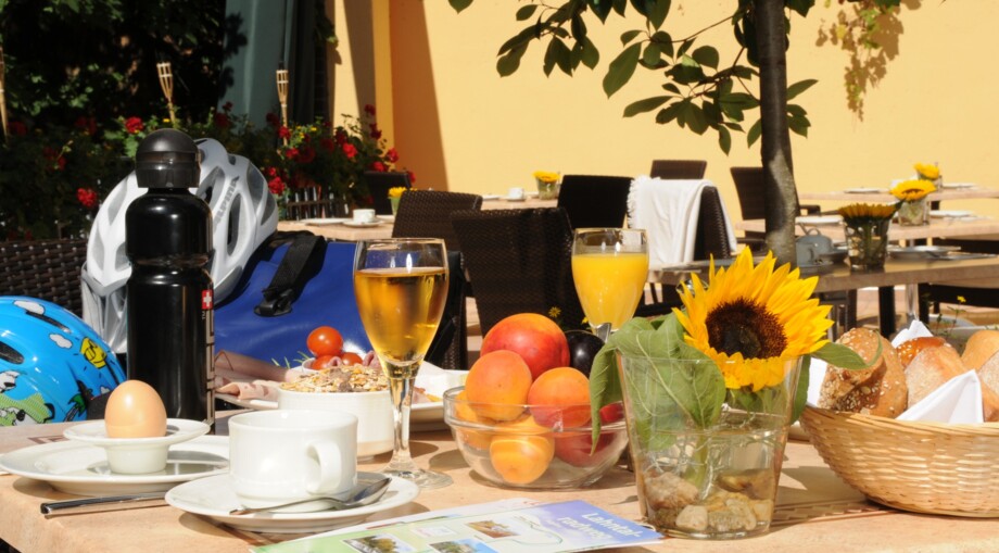 Outdoor breakfast setting with fresh fruit, orange juice, coffee, bread basket, and sunflower on a sunny terrace with garden seating