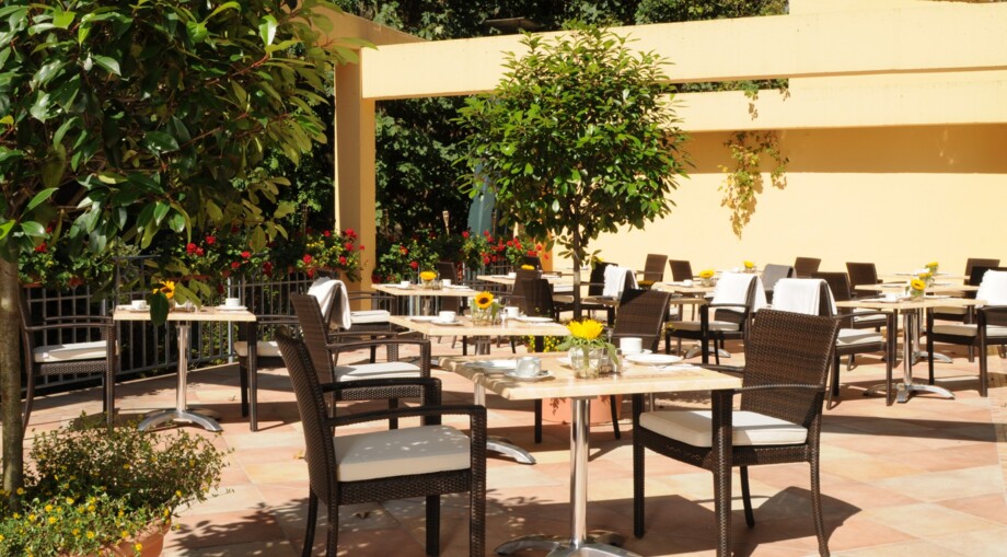 Sunny outdoor hotel terrace with wicker chairs, marble tables, sunflowers, and lush greenery for relaxed dining