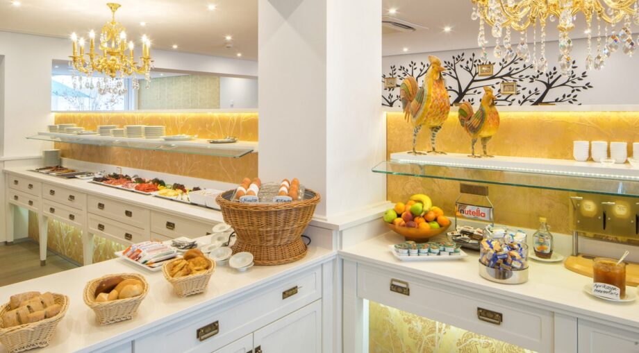 Bright breakfast buffet with fresh bread baskets, boiled eggs, fruit bowl, jams, and cold cuts under elegant chandeliers in a cozy dining area.