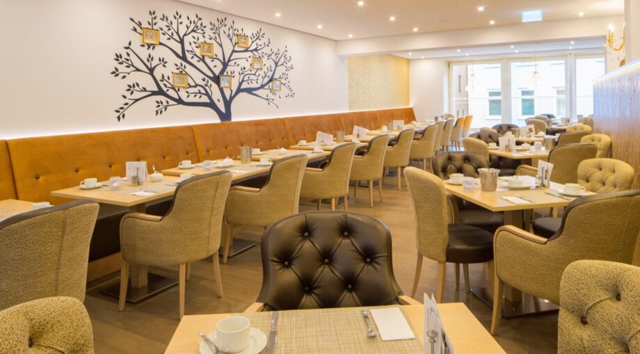 Bright hotel dining room with tufted chairs, wooden tables set for breakfast, and a decorative tree mural on the wall