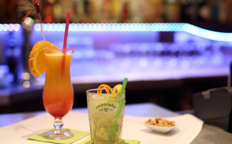 Zwei farbenfrohe Cocktails mit Fruchtgarnitur auf einer Bar, Schale mit Nüssen und blauem LED-Licht im Hintergrund