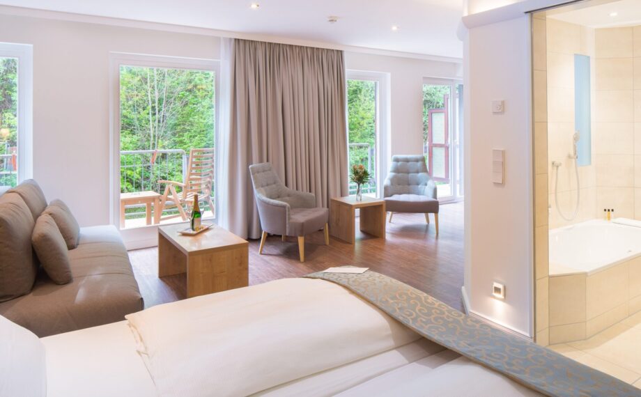 Helle Hotelsuite mit Kingsize-Bett, gemütlicher Sitzecke, Balkon mit Blick auf grüne Landschaft und modernem Bad mit Badewanne