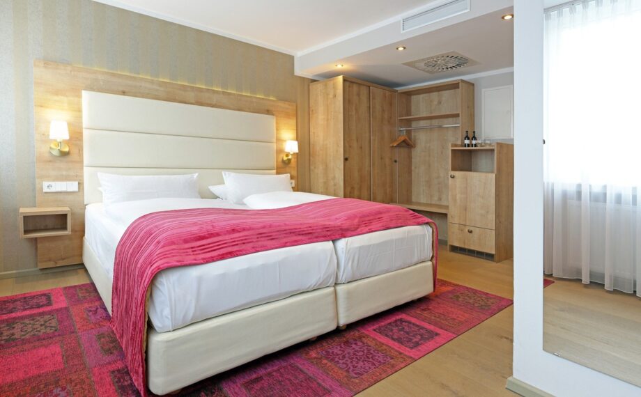 Modernes Hotelzimmer mit zwei zusammengestellten Betten, pinker Tagesdecke, Holzkleiderschrank, weicher Beleuchtung und großem Fenster mit Gardinen