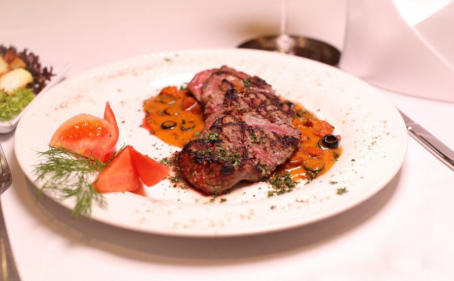 Gegrillte Steakstreifen mit Kräutergarnitur und würziger Tomaten-Oliven-Sauce, dazu frische Tomatenecken und Dill auf weißem Teller